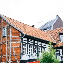 Urokliwy dom szachulcowy z ceglanymi ścianami i białymi belkami, otoczony innymi budynkami i zielenią.