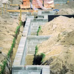 Budowa fundamentów z bloczków betonowych, z widocznymi wykopami i usypiskami ziemi, uwzględniającymi strefy przemarzania gruntu.