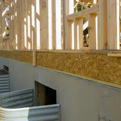 Budowa domu w stanie surowym. Widoczny betonowy fundament, płyta OSB jako izolacja i drewniana konstrukcja ścian. Widać też okna piwniczne.