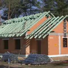 Budowa domu w toku, widoczna konstrukcja dachu i ściany z cegły. Materiały budowlane leżą na ziemi. To przykład metody gospodarczej.