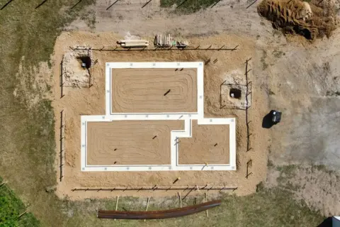 Widok z lotu ptaka na plac budowy z fundamentami. Różne rodzaje fundamentów są widoczne, w tym ławy fundamentowe i wykopy pod słupy.