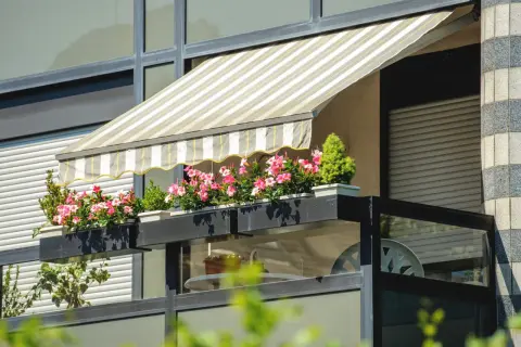 Zadaszenia balkonowe w paski chronią kwitnące pelargonie. Balkon z roślinnością i meblami.