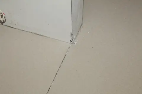 Widok na narożnik ściany i podłogi, gdzie widać, jak zrobić dylatacje w wylewce. Widoczne są szczeliny i fragmenty materiału.