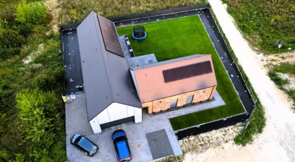 Nowoczesny dom pod klucz, gotowy do zamieszkania. Dwa samochody na podjeździe, panele słoneczne na dachu, zadbany ogród z trampoliną.