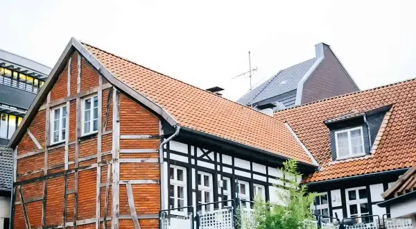 Urokliwy dom szachulcowy z ceglanymi ścianami i białymi belkami, otoczony innymi budynkami i zielenią.