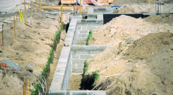 Budowa fundamentów z bloczków betonowych, z widocznymi wykopami i usypiskami ziemi, uwzględniającymi strefy przemarzania gruntu.