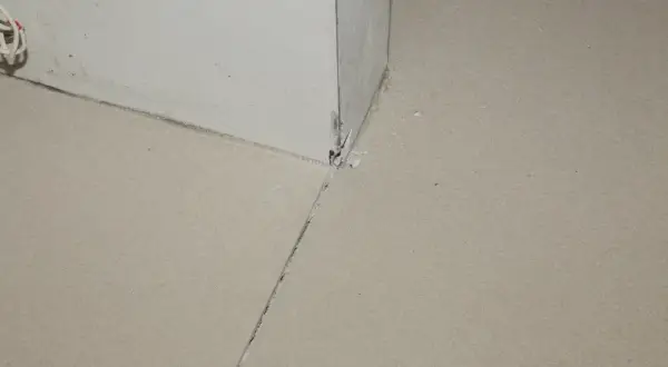 Widok na narożnik ściany i podłogi, gdzie widać, jak zrobić dylatacje w wylewce. Widoczne są szczeliny i fragmenty materiału.
