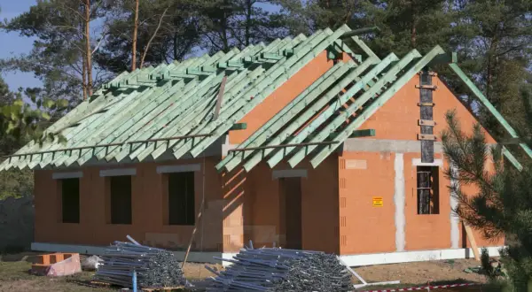 Budowa domu w toku, widoczna konstrukcja dachu i ściany z cegły. Materiały budowlane leżą na ziemi. To przykład metody gospodarczej.