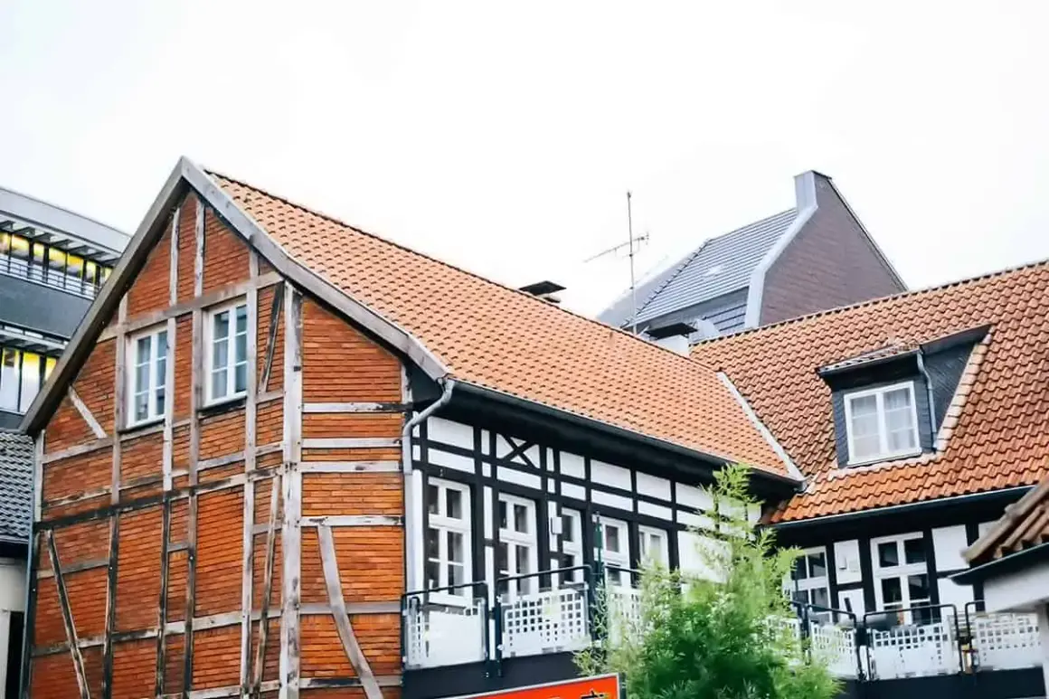 Urokliwy dom szachulcowy z ceglanymi ścianami i białymi belkami, otoczony innymi budynkami i zielenią.