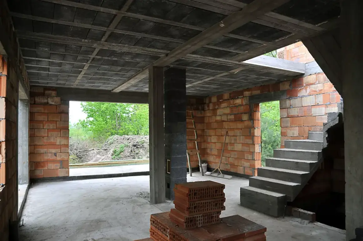 Budowa domu w stanie surowym. Widać betonowy strop typu ackerman, ściany z pustaków ceramicznych, schody i materiały budowlane.