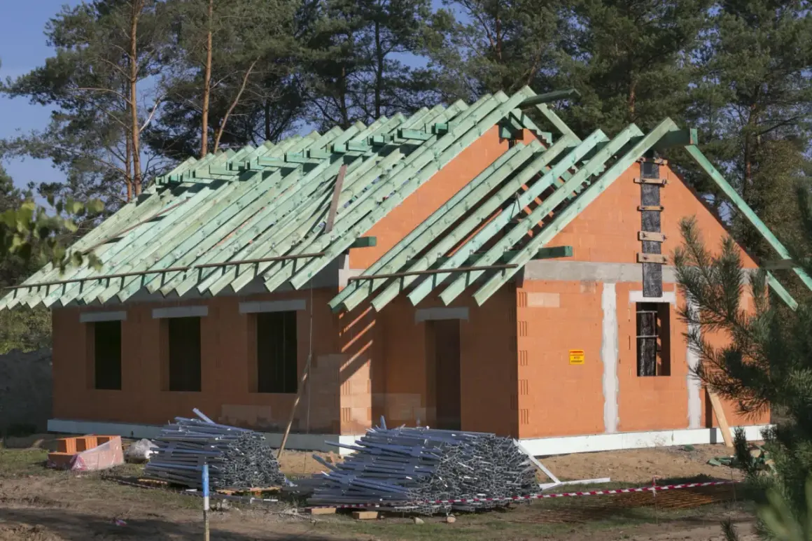 Budowa domu w toku, widoczna konstrukcja dachu i ściany z cegły. Materiały budowlane leżą na ziemi. To przykład metody gospodarczej.