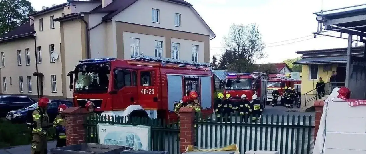 Strażacy w akcji po pożarze fotowoltaiki. Czerwone wozy strażackie i mundurowi przy budynku.