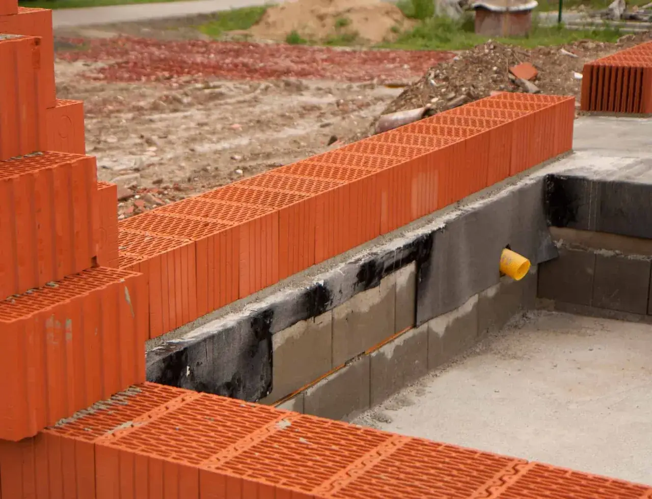 Budowa domu w toku. Widoczne pomarańczowe pustaki ceramiczne, fundamenty i żółta rura kanalizacyjna. Trwa ustalanie, ile pustaków na m2 będzie potrzebne.