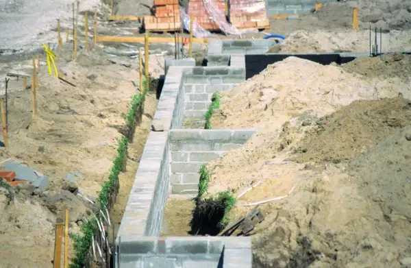 Budowa fundamentów z bloczków betonowych, z widocznymi wykopami i usypiskami ziemi, uwzględniającymi strefy przemarzania gruntu.