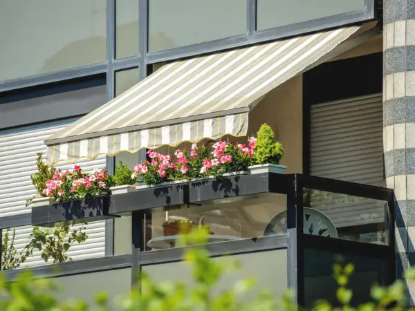 Zadaszenia balkonowe w paski chronią kwitnące pelargonie. Balkon z roślinnością i meblami.