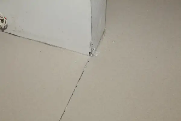 Widok na narożnik ściany i podłogi, gdzie widać, jak zrobić dylatacje w wylewce. Widoczne są szczeliny i fragmenty materiału.